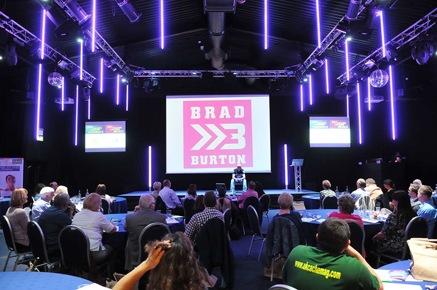 Wide view of a Brad Burton keynote with the BB logo on a giant screen and purple light tubes hanging from the rig