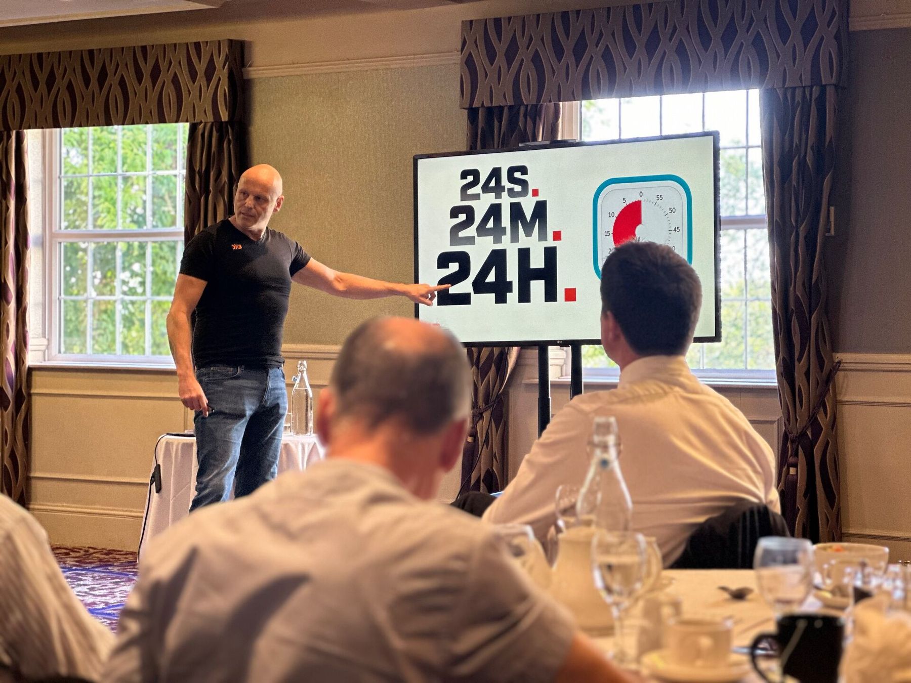 Brad Burton mid-keynote pointing at his '24S. 24M. 24H.' slide with the Time Timer beside it, audience in the foreground
