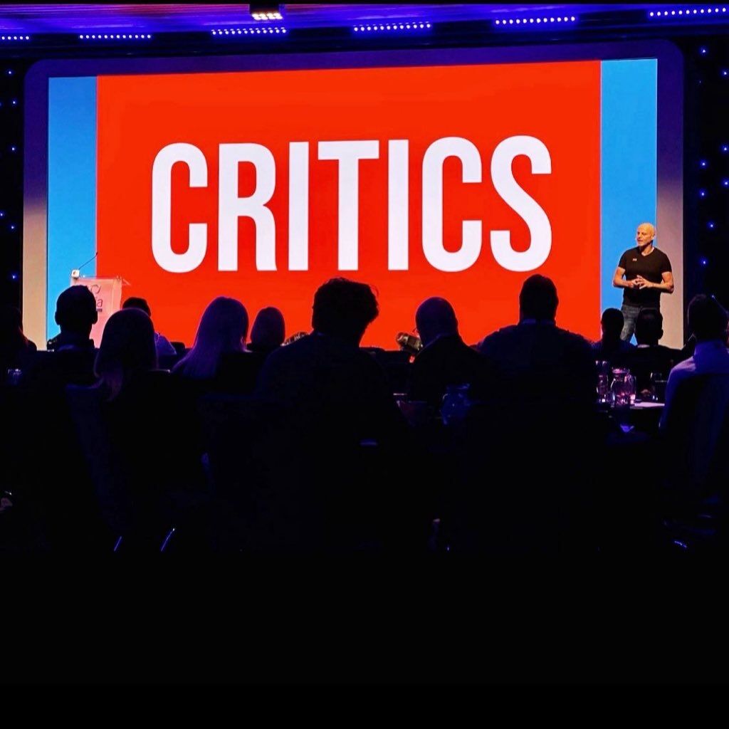 Brad on stage in front of a giant orange 'CRITICS' slide, audience in silhouette