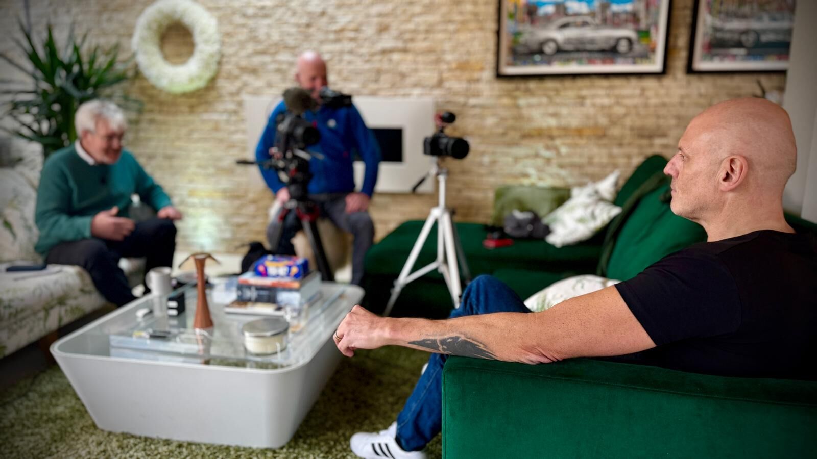 Brad Burton being interviewed on a green sofa at home, with a film crew and two cameras across the coffee table