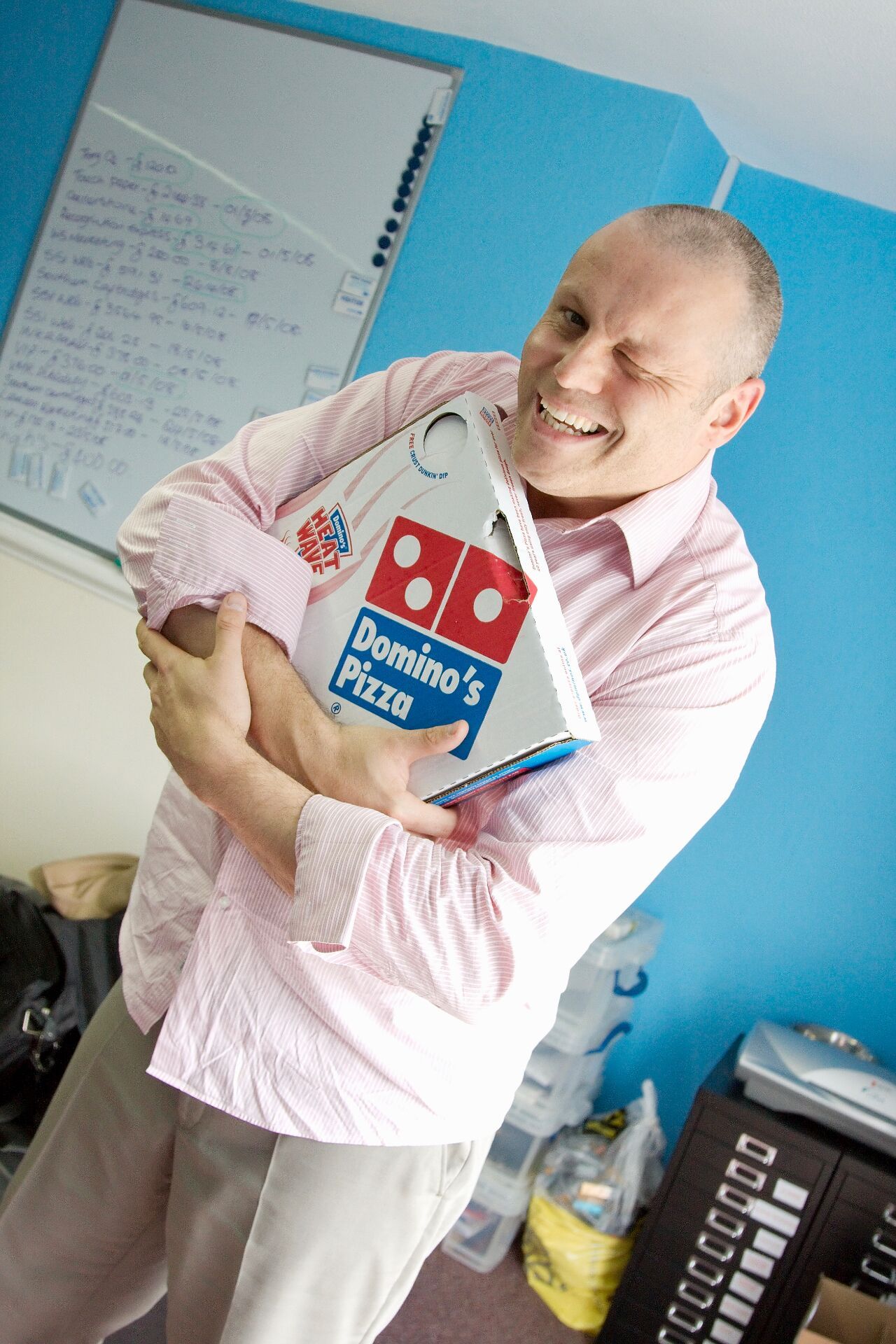 A young Brad Burton in his old office hugging a Domino's pizza box, grinning, the whiteboard behind him covered in handwritten debt totals and dates