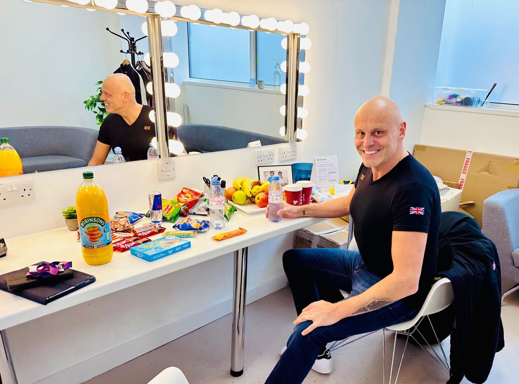 Brad Burton sitting in a dressing room with a vanity-bulb mirror behind him and a snack table covered in fruit, sweets, water and Costa coffees before going on stage