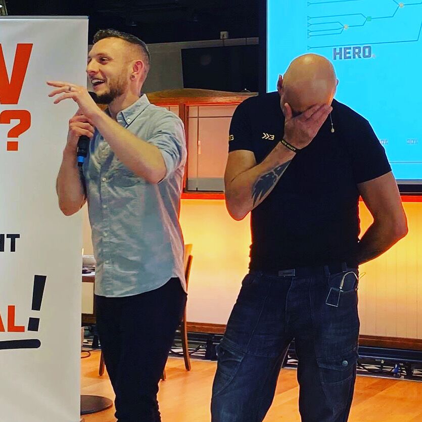 Brad on stage in stitches with the host beside him, mic in hand