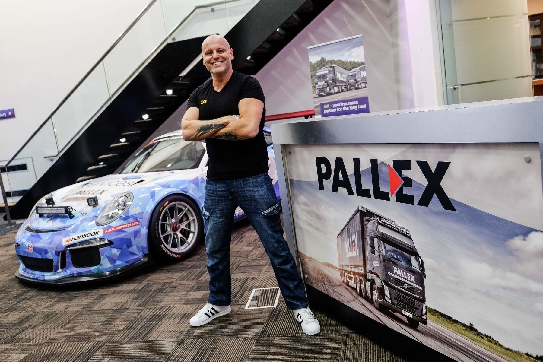 Brad Burton on a brand shoot at Pall-Ex HQ, arms folded, beside the Pall-Ex Porsche racing car