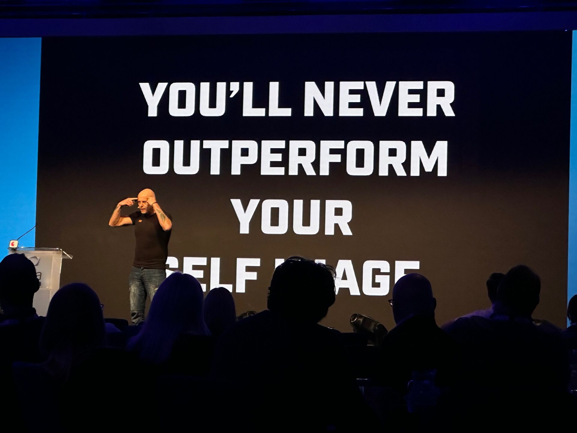 Brad on a fully lit main stage in front of a giant slide reading 'YOU'LL NEVER OUTPERFORM YOUR SELF IMAGE'
