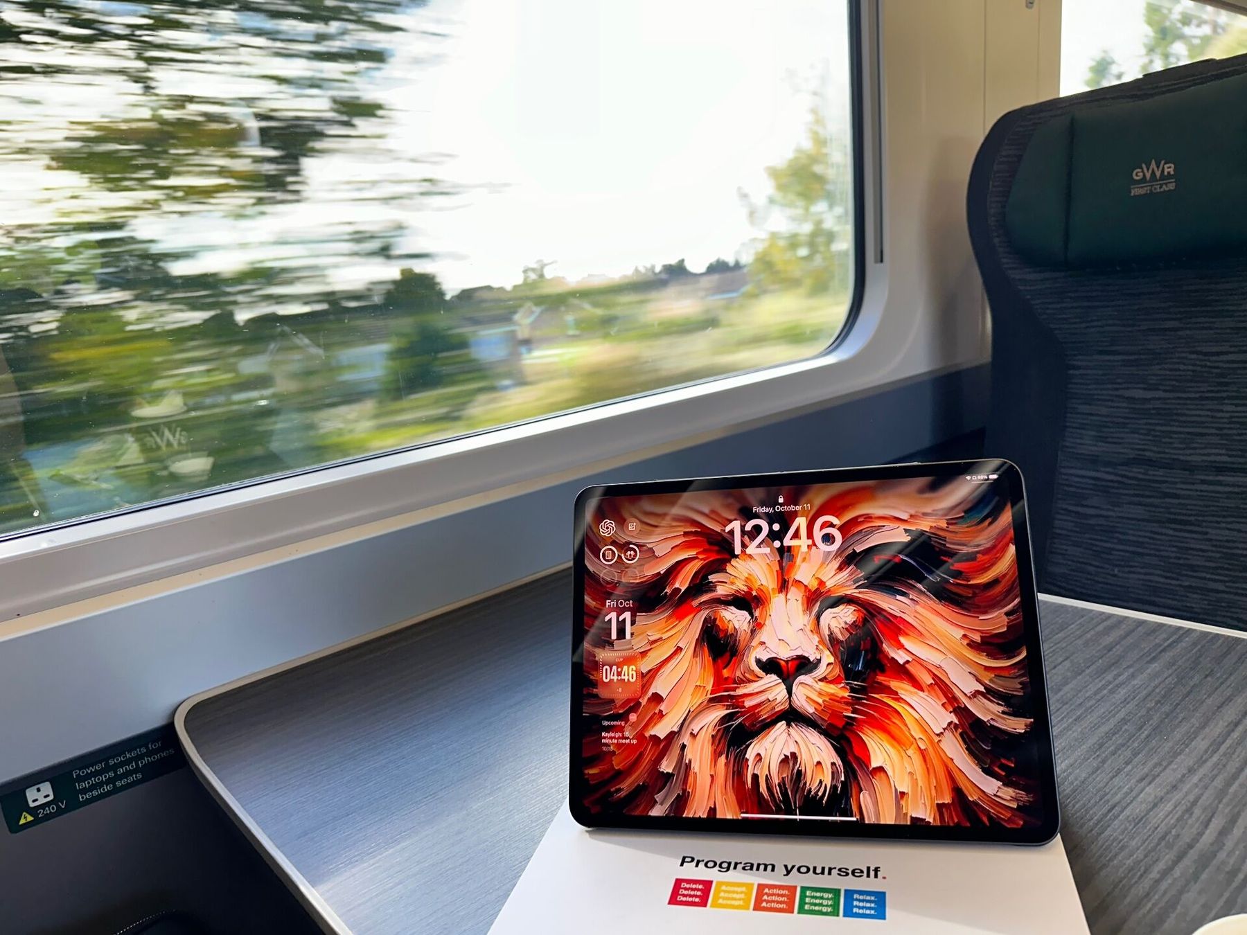 iPad on a GWR First Class table showing a fiery lion lock screen, with a 'Program yourself' card underneath as the train passes green countryside